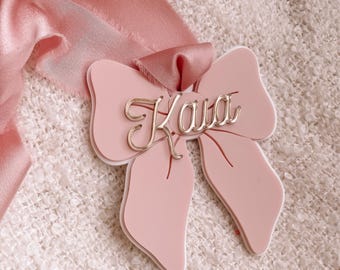 May include: A light pink bow ornament featuring a layered design with a white backing. The name Kaia is written in a metallic gold cursive script across the center. A matching pink silk ribbon with frayed edges is attached to the top of the bow.