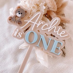 May include: A beige cake topper featuring a brown teddy bear icon and the raised white and light blue text "Archie is ONE". The topper has a long, thin beige stem extending from the bottom of the main plaque, which is shaped with rounded edges.