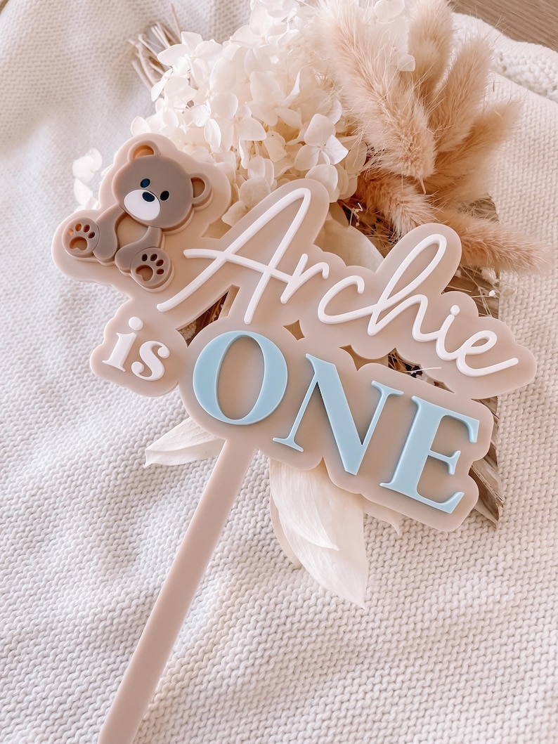 May include: A beige cake topper featuring a brown teddy bear icon and the raised white and light blue text "Archie is ONE". The topper has a long, thin beige stem extending from the bottom of the main plaque, which is shaped with rounded edges.