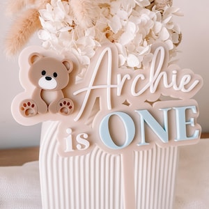 May include: A beige cake topper featuring a brown bear illustration on the left side. The text reads Archie in white script, followed by is in white, and ONE in light blue block letters. The topper has a long stem for insertion into a cake.