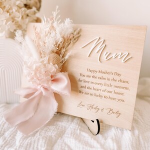 May include: A wooden plaque with the word "Mum" in white script, adorned with dried flowers and a pink ribbon. The plaque features a Mother's Day message and is supported by a black stand.