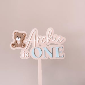 May include: A cake topper featuring a light pink base with a brown teddy bear icon on the left. The text reads "Archie" in white script, with "is ONE" in light blue block letters below. The topper includes a vertical stake for insertion.