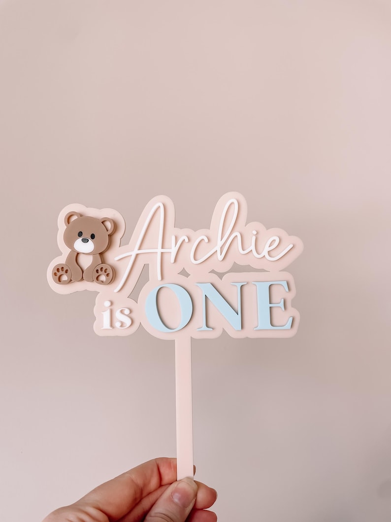 May include: A cake topper featuring a light pink base with a brown teddy bear icon on the left. The text reads "Archie" in white script, with "is ONE" in light blue block letters below. The topper includes a vertical stake for insertion.