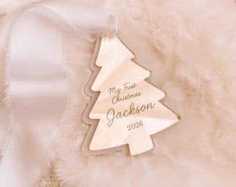 May include: A reflective, tree-shaped Christmas ornament featuring the engraved text "My First Christmas Jackson 2026" in a script and sans-serif font. The ornament is attached to a wide, white satin ribbon with frayed edges for hanging purposes.