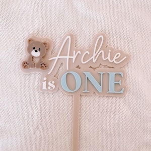 May include: A beige acrylic cake topper featuring a brown bear icon on the left side. The text reads "Archie is ONE" in a combination of white script and blue block lettering. The topper includes a single pointed stake for insertion into a cake.