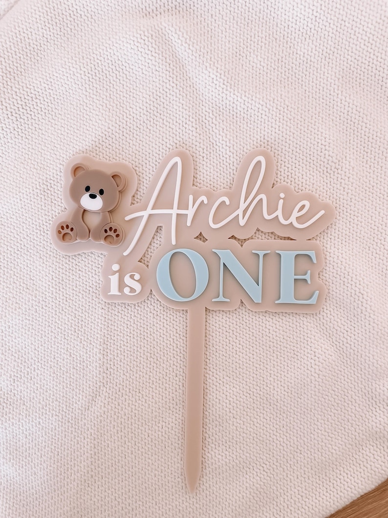 May include: A beige acrylic cake topper featuring a brown bear icon on the left side. The text reads "Archie is ONE" in a combination of white script and blue block lettering. The topper includes a single pointed stake for insertion into a cake.