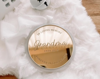 May include: A circular memorial ornament with a reflective gold mirror finish. The text reads IN LOVING MEMORY, Grandma, 1934-2015, and FOREVER IN OUR HEARTS. The ornament has a smooth, light-coloured border and a white ribbon for hanging purposes.