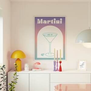 May include: A framed print with the word "Martini" above a teal line drawing of a martini glass. A yellow lamp, a small pink lamp, and a multi-colored candle holder sit on a white cabinet.