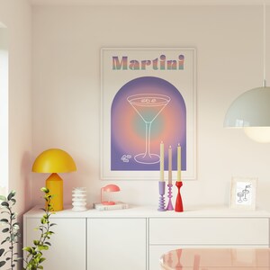 Op de afbeelding: Een ingelijste print met het woord "Martini" in pastelkleuren boven een lijntekening van een martiniglas. De print wordt weergegeven boven een witte kast met een gele lamp, kaarsen en andere decoratieve items.