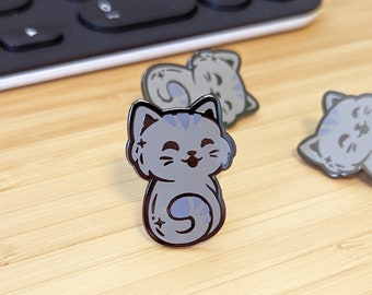 May include: Three gray cat enamel pins with black outlines and light blue stripes. The pins feature a smiling cat design, with one pin standing upright and two lying down. The pins are on a light wood surface.