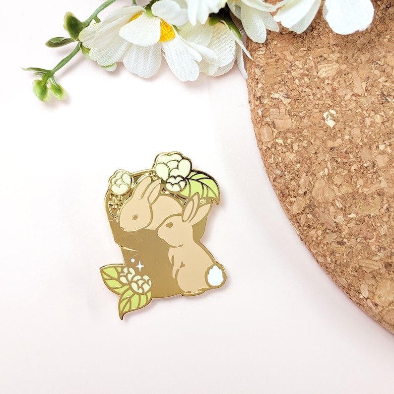 May include: A gold-colored enamel pin featuring two tan rabbits nestled in a basket with white flowers and green leaves. The pin has a shiny finish and is set against a light pink background.