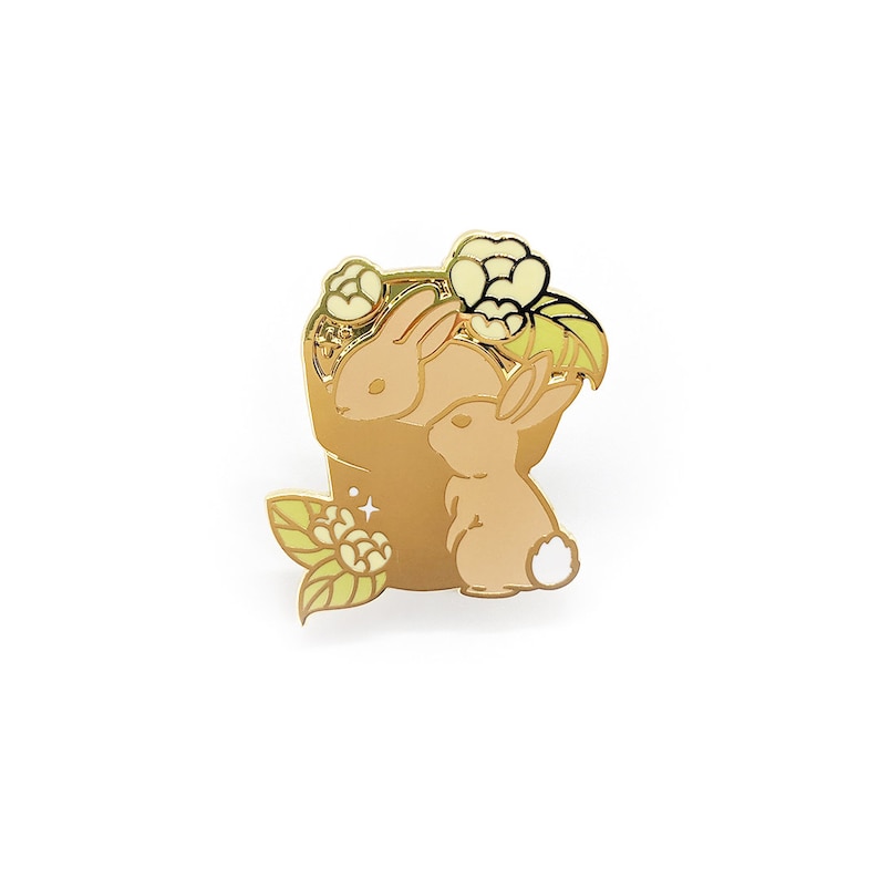 May include: A gold-colored enamel pin featuring two tan rabbits nestled in a pot with cream-colored flowers and green leaves. The pin has a shiny finish and is set against a white background. The design is detailed and whimsical.