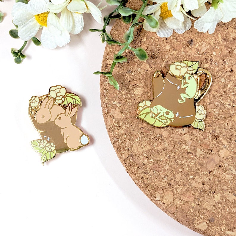 May include: Two enamel pins. One pin features two brown bunnies in a basket with flowers. The other pin is a brown teapot with a frog and flowers. Both pins have gold accents and light green and white details.