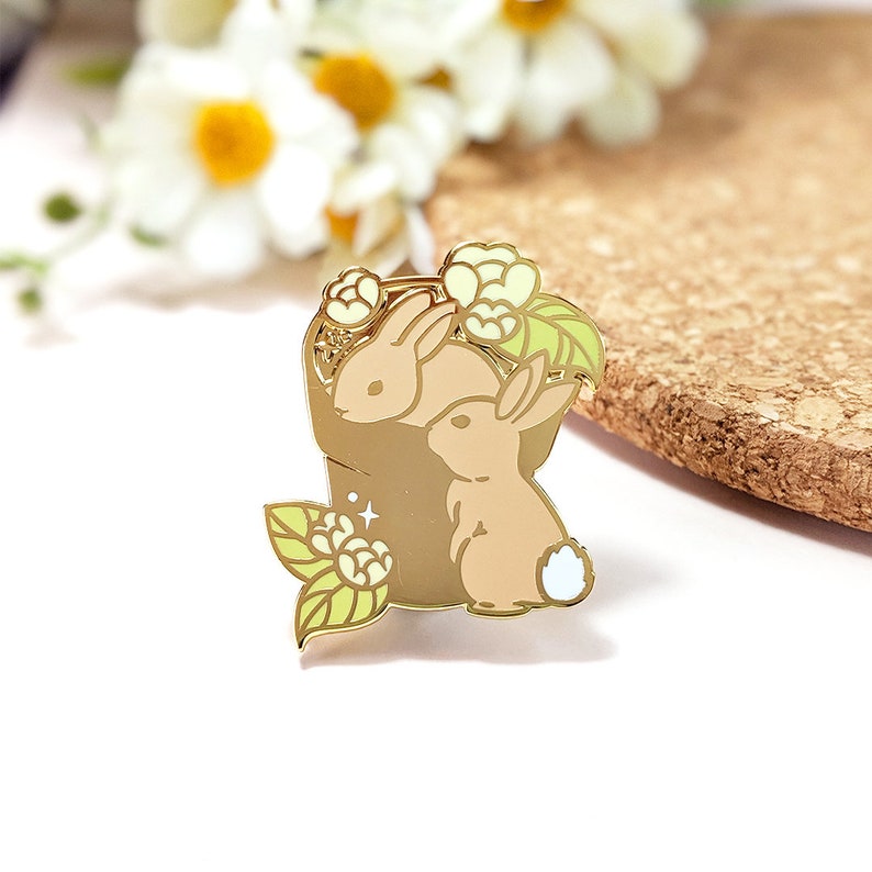 May include: A gold-colored enamel pin featuring two tan rabbits nestled in a brown basket with white flowers and green leaves. The pin has a white background and is a decorative accessory.