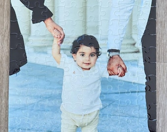 Può includere: Un puzzle fotografico di famiglia con un bambino piccolo che tiene le mani di due adulti. Il bambino indossa una camicia bianca e pantaloni beige, mentre gli adulti indossano abiti casual. I pezzi del puzzle sono disposti in modo casuale, creando un'esperienza divertente e stimolante.