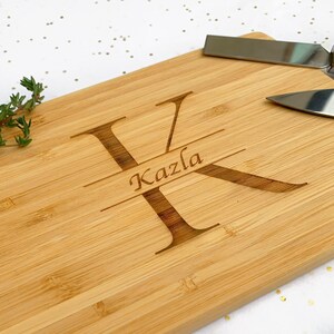 Op de afbeelding: Een gepersonaliseerd bamboe snijplank met de letter "K" en de naam "Kazla" erop gegraveerd. De plank is lichtbruin en heeft een natuurlijke houtnerf.