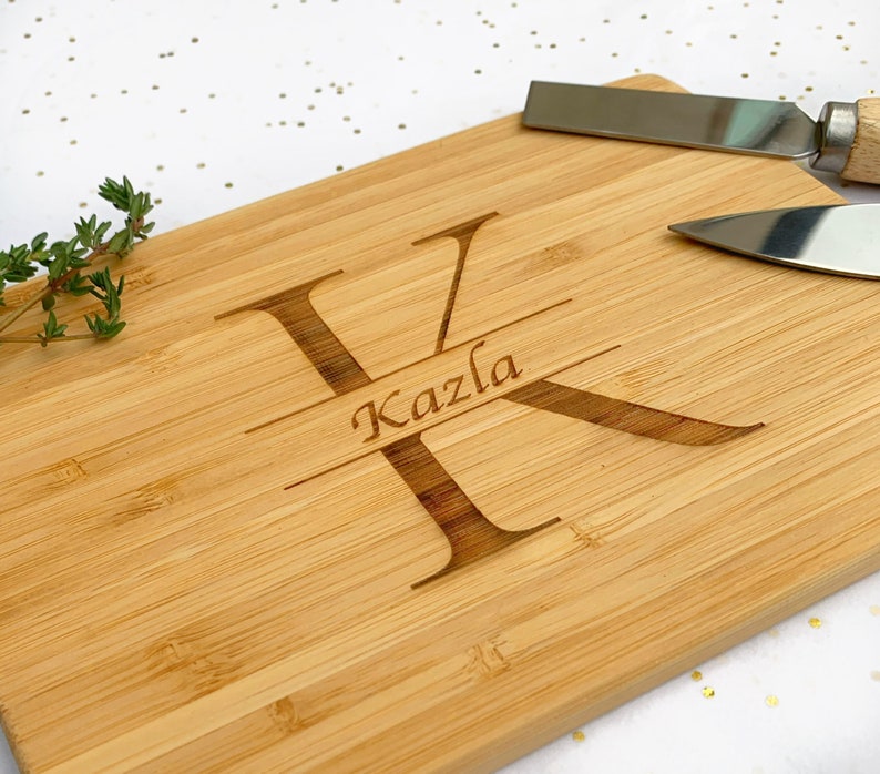 Op de afbeelding: Een gepersonaliseerd bamboe snijplank met de letter "K" en de naam "Kazla" erop gegraveerd. De plank is lichtbruin en heeft een natuurlijke houtnerf.