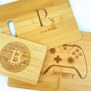 Op de afbeelding: Drie houten snijplanken met gepersonaliseerde gravures. De eerste plank heeft de letter "P" en de tekst "Ps Coot Board". De tweede plank heeft een Bitcoin-logo en de tekst "TROY OZ 999 FINE COPPER BITCOIN DIGITAL DECENTRALIZED PEER TO PEER METALS MONETARY". De derde plank heeft een videogamecontroller en de tekst "Jeremy".
