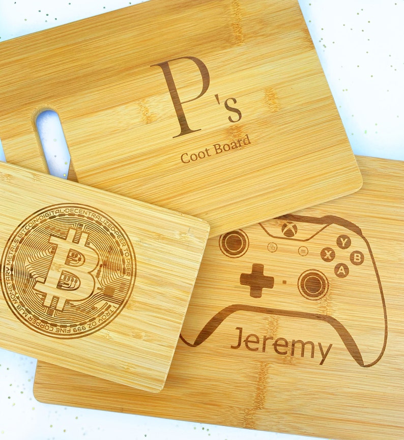 Op de afbeelding: Drie houten snijplanken met gepersonaliseerde gravures. De eerste plank heeft de letter "P" en de tekst "Ps Coot Board". De tweede plank heeft een Bitcoin-logo en de tekst "TROY OZ 999 FINE COPPER BITCOIN DIGITAL DECENTRALIZED PEER TO PEER METALS MONETARY". De derde plank heeft een videogamecontroller en de tekst "Jeremy".