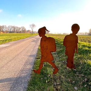 Può includere: Due sagome metalliche arrugginite di bambini che camminano, una con uno zaino, su un campo erboso accanto a una strada asfaltata.