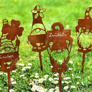 Op de afbeelding: Vier roestige metalen tuinmarkeringen in de vorm van dwergen met handgeschreven labels. De labels luiden "Tomaten", "Zwiebeln" en twee onleesbare labels.