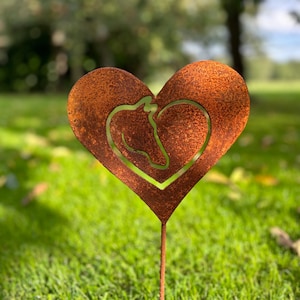 Könnte beinhalten: Ein rostiger, herzförmiger Gartenpfahl aus Metall mit einer Aussparung für einen Pferdekopf. Der Pfahl ist in grünem Gras gesteckt.