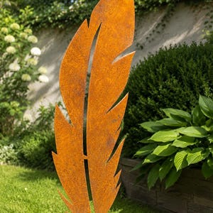Op de afbeelding: Een grote, verweerde metalen veersculptuur met een roestige oranje afwerking staat in een tuin. De veer heeft een gedetailleerd uitgesneden ontwerp en is geplaatst in een grasveld met weelderig groen op de achtergrond. Een decoratief tuinaccessoire.