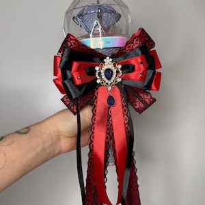Pode incluir: Um bastão de luz decorativo com um topo de globo transparente e facetado. O bastão de luz é adornado com um grande laço vermelho e preto, embelezado com renda e uma peça central com joias. O texto "HOSHI" é visível na base.