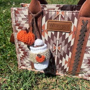 May include: A brown and white patterned tote bag with brown leather handles and accents. A small, crocheted pumpkin and a crocheted coffee cup charm with a pumpkin accent hang from the handle. The bag has a leather tag that says "Wrangler".