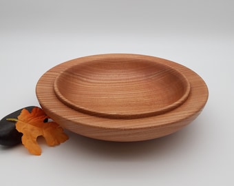 May include: Two wooden bowls, one nested inside the other, are made of light brown wood with a grain pattern. The bowls are round and have a slight curve to them.