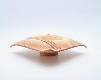 May include: A light-colored wooden bowl with a wide, undulating rim and a circular base. The bowl has a central depression and is adorned with three dark brown stripes. The bowl is likely handmade and has a natural, organic aesthetic.