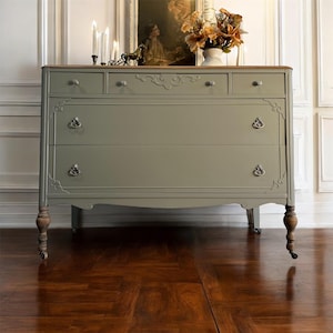 May include: A vintage dresser with a light green paint finish and wooden legs. The dresser has five drawers with ornate carvings and silver handles. The top drawer is a lighter wood color.