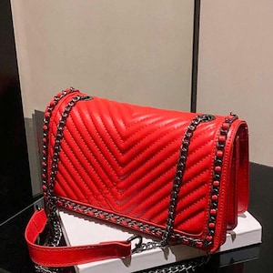 May include: A vibrant red handbag with a quilted chevron pattern. The bag features a silver chain strap and trim. The design includes a top handle and a long shoulder strap. The bag is sitting on a white surface.