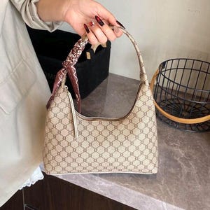 Può includere: Borsa a tracolla beige con un motivo geometrico ripetuto. La borsa ha una forma curva, un manico superiore e una sciarpa decorativa legata al manico. La borsa è tenuta da una persona.