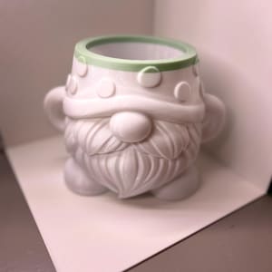 May include: A white gnome-shaped planter with a light green rim. The gnome features a textured beard, a round nose, and a hat adorned with raised circular polka dots. The planter has two small handles on the sides and a wide, open top opening.