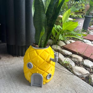 May include: A bright yellow pineapple-shaped planter featuring a gray arched door with a circular emblem, two small gray circular windows, and a gray pipe protrusion on the side. A tall, green, variegated snake plant grows out of the top opening.