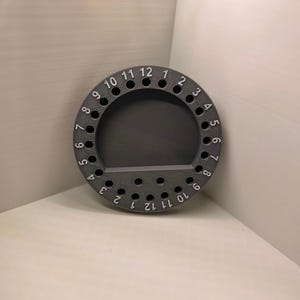 May include: A circular dark gray organizer tray featuring a series of small, evenly spaced holes along the perimeter. Each hole is labeled with a white printed number, with two separate sequences of numbers from 1 to 12 arranged around the rim.