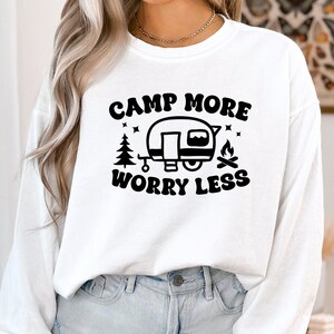May include: White long-sleeved shirt with black text that reads "Camp More Worry Less" and a black illustration of a camper, a tree, and a campfire.