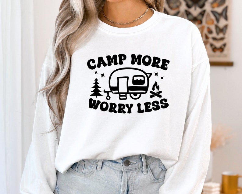May include: White long-sleeved shirt with black text that reads "Camp More Worry Less" and a black illustration of a camper, a tree, and a campfire.