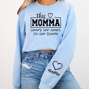 May include: Light blue sweatshirt with the text "this MOMMA wears her heart on her sleeve." A black heart and the word "name" are on the sleeve. The sweatshirt is made of a soft material.