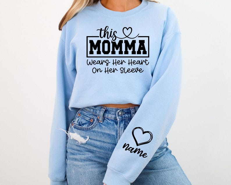 May include: Light blue sweatshirt with the text "this MOMMA wears her heart on her sleeve." A black heart and the word "name" are on the sleeve. The sweatshirt is made of a soft material.
