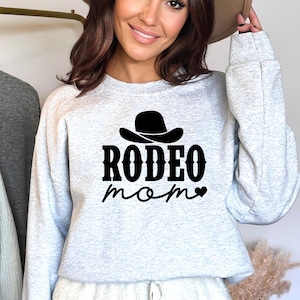 Op de afbeelding: Grijs sweatshirt met een zwarte cowboyhoed-afbeelding en de tekst "Rodeo Mom" met een klein hartje.