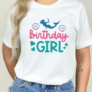 May include: White t-shirt featuring a mermaid graphic, seashells, and the text "Birthday Girl" in pink and teal.