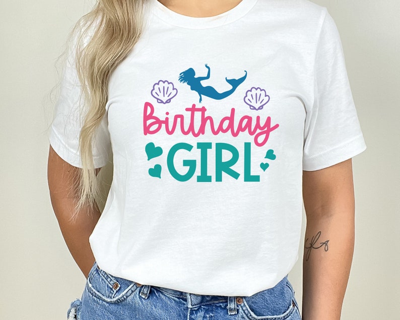 May include: White t-shirt featuring a mermaid graphic, seashells, and the text "Birthday Girl" in pink and teal.