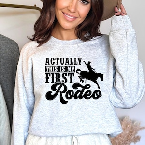 Op de afbeelding: Een grijs sweatshirt met de tekst "Actually this is my first rodeo" in zwarte letters met een silhouet van een cowgirl die een paard berijdt.