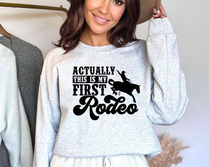 Op de afbeelding: Een grijs sweatshirt met de tekst "Actually this is my first rodeo" in zwarte letters met een silhouet van een cowgirl die een paard berijdt.