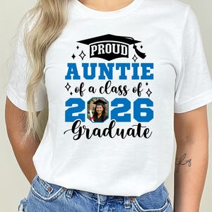 Peut inclure: T-shirt blanc avec l'inscription "PROUD AUNTIE of a class of 2026 Graduate" en bleu et noir. Le motif comprend un chapeau de graduation et une photo d'un dipl&ocirc;m&eacute;. Un tee-shirt d&eacute;contract&eacute;.