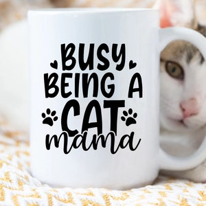 Pu&ograve; includere: Tazza in ceramica bianca con testo nero che recita "Busy Being a Cat mama" con impronte di zampe su entrambi i lati della parola "Cat". Un gatto bianco con macchie marroni e nere spunta da dietro la tazza.
