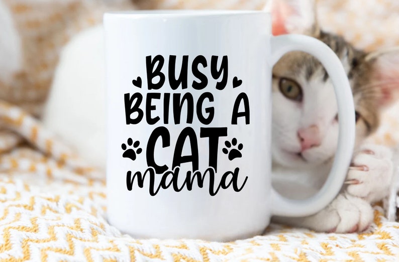 Pu&ograve; includere: Tazza in ceramica bianca con testo nero che recita "Busy Being a Cat mama" con impronte di zampe su entrambi i lati della parola "Cat". Un gatto bianco con macchie marroni e nere spunta da dietro la tazza.