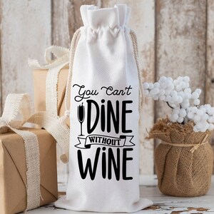 May include: White cotton wine bag with black text that reads "You Can't Dine Without Wine". The bag has a drawstring closure and is sitting on a wooden surface.